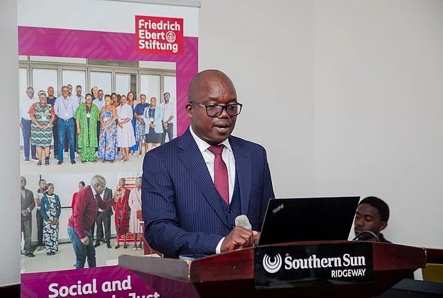 Black man in suit speaking at Southern Sun Ridgeway podium with Friedrich Ebert Stiftung banner in background.