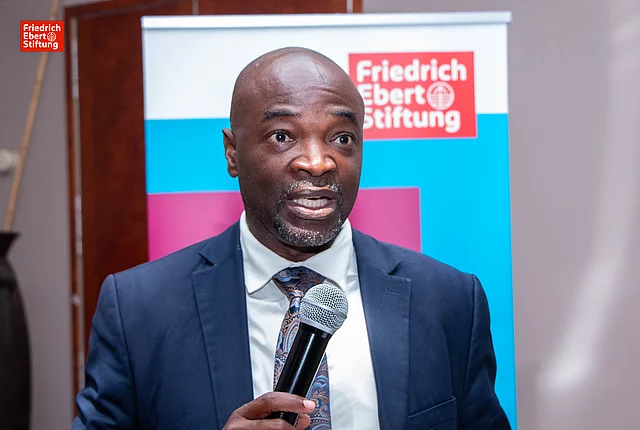 An elderly African man in a dark suit and patterned tie speaks into a microphone at an event. He stands in front of a banner featuring the Friedrich Ebert Stiftung logo, with a blue and red background. The setting appears formal.