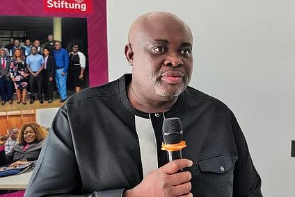 Black man in black and white shirt holding a microphone, with a group photo and a seated person visible in the background.