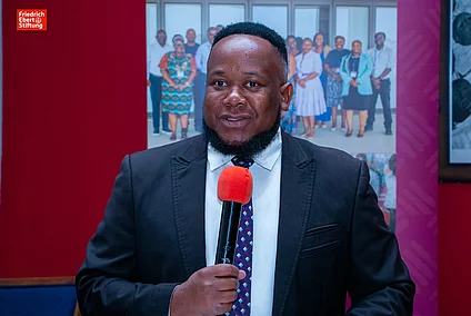 Black man in suit holding a red microphone, standing indoors with a group photo and Friedrich Ebert Stiftung logo in the background