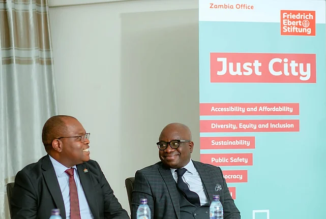 Two African men in suits sit at a table in front of a banner that reads "Just City" and lists themes such as "Accessibility and Affordability," "Diversity, Equity and Inclusion," "Sustainability," "Public Safety," and "Democracy" alongside the Friedrich Ebert Stiftung logo.