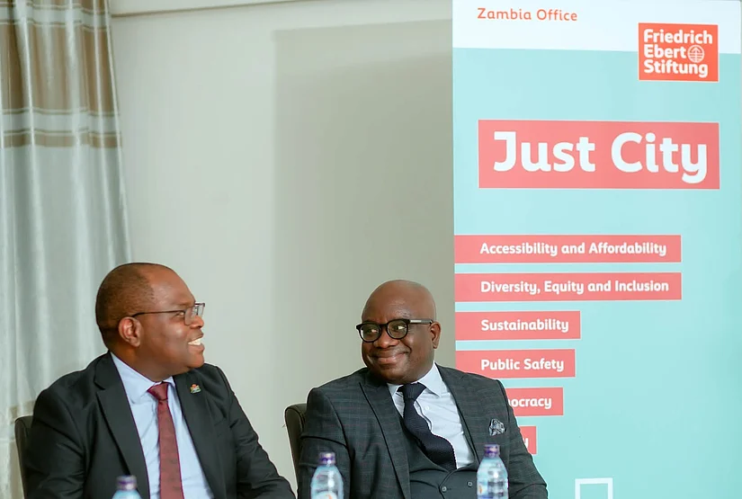 Two African men in suits sit at a table in front of a banner that reads "Just City" and lists themes such as "Accessibility and Affordability," "Diversity, Equity and Inclusion," "Sustainability," "Public Safety," and "Democracy" alongside the Friedrich Ebert Stiftung logo.