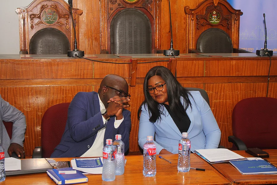 Zambian Kitwe mayor with a male Zambian in the council chambers, engaged in conversation. Both wear glasses and blue blazers. Water bottles and papers are on the table.
