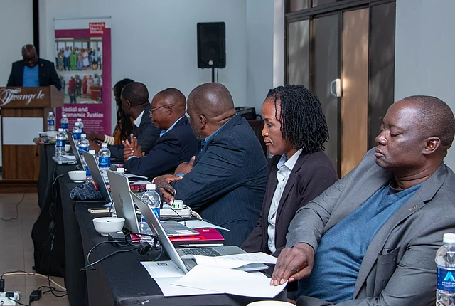 People seated at a conference table with laptops and documents, listening to a speaker at a podium in a meeting room.