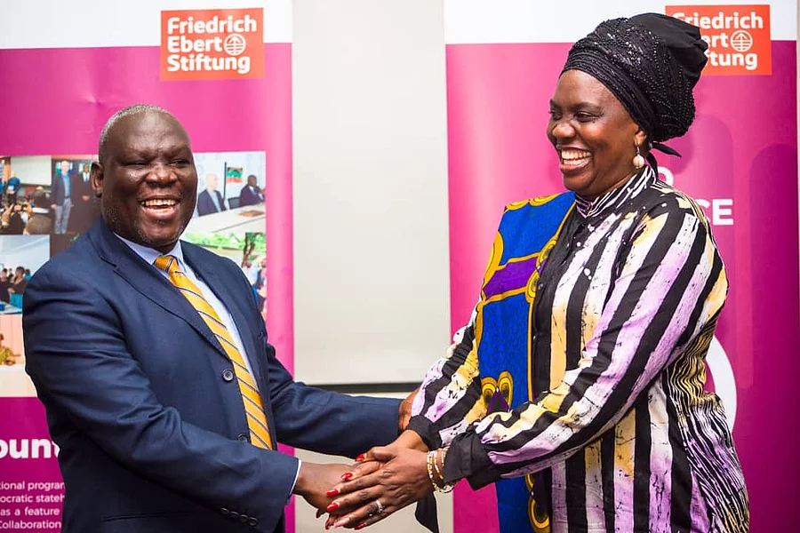 Two Zambian individuals, a man in a suit and a woman in colorful attire, are shaking hands and smiling warmly. They stand in front of pink banners.