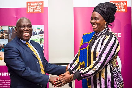 Two Zambian individuals, a man in a suit and a woman in colorful attire, are shaking hands and smiling warmly. They stand in front of pink banners.
