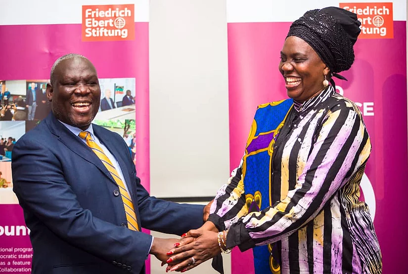 Two Zambian individuals, a man in a suit and a woman in colorful attire, are shaking hands and smiling warmly. They stand in front of pink banners.