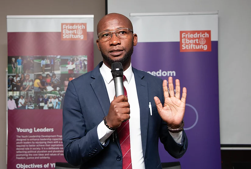 A middle-aged African man in a suit and red tie holds a microphone and raises his hand while speaking at an event with Friedrich Ebert Stiftung banners in the background.