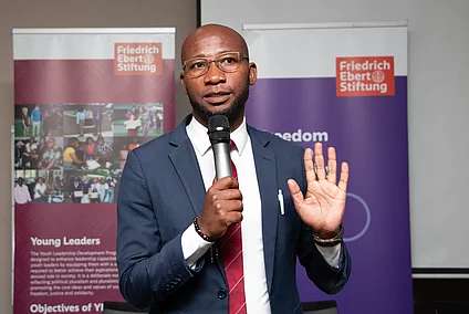 A middle-aged African man in a suit and red tie holds a microphone and raises his hand while speaking at an event with Friedrich Ebert Stiftung banners in the background.