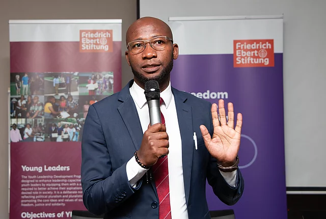 A middle-aged African man in a suit and red tie holds a microphone and raises his hand while speaking at an event with Friedrich Ebert Stiftung banners in the background.