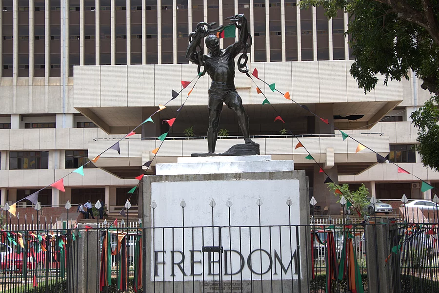 Zambian Freedom statue of a man breaking chains stands before a building, adorned with colorful triangular flags. The pedestal reads "FREEDOM," symbolizing liberation.