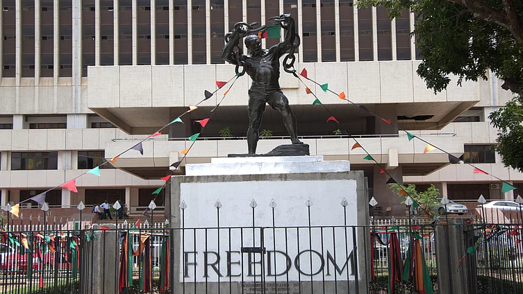 Zambian Freedom statue of a man breaking chains stands before a building, adorned with colorful triangular flags. The pedestal reads "FREEDOM," symbolizing liberation.