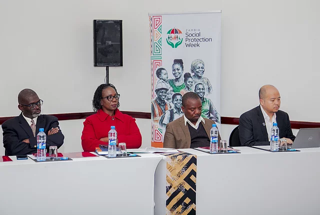 Four people sit at a conference table with water bottles, papers, and a laptop; a "Zambia Social Protection Week" banner is displayed behind them.