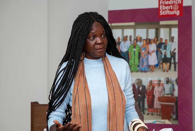 Black woman with long braided hair and orange scarf speaking in front of a Friedrich Ebert Stiftung banner featuring group photos.