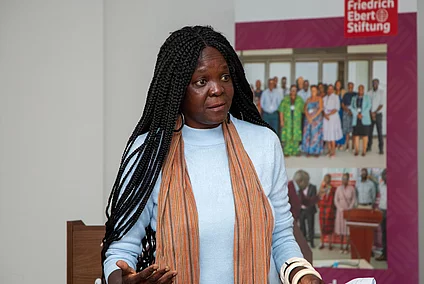 Black woman with long braided hair and orange scarf speaking in front of a Friedrich Ebert Stiftung banner featuring group photos.