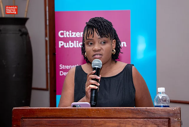 A middle-aged African woman with short, curly hair stands at a podium holding a microphone, speaking to an audience. She wears a black sleeveless dress and appears focused. A water bottle is on the podium, and a colorful backdrop featuring text related to public participation is visible behind her.