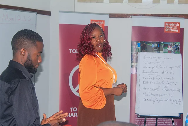 Two people in a workshop setting; one speaker in orange, flip chart with handwritten notes and Friedrich Ebert Stiftung banners in the background.