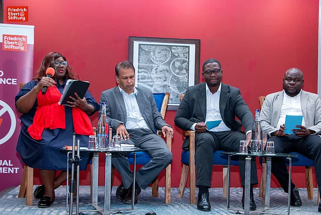 Panel discussion with four speakers seated, one holding a microphone, against a red wall with Friedrich Ebert Stiftung branding.
