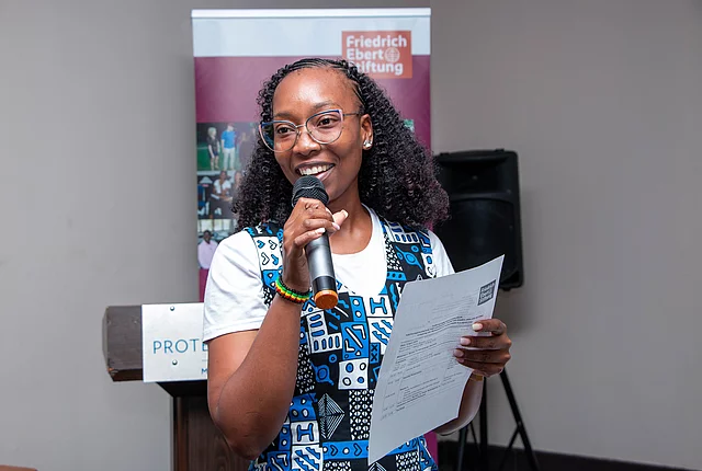 A middle-aged African woman holding a microphone and a sheet of paper speaks at an indoor event, standing in front of a podium and a Friedrich Ebert Stiftung banner.