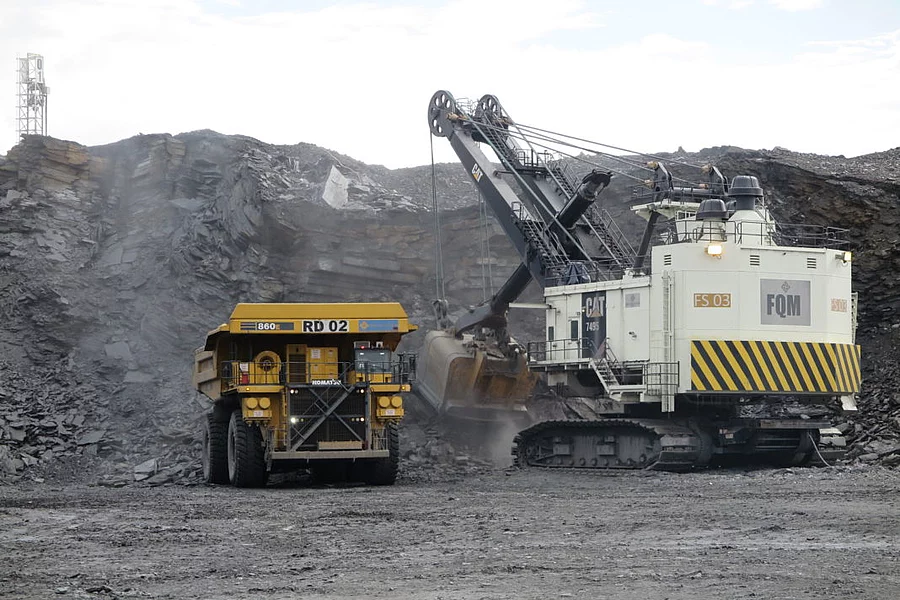 A large excavator loads rock into a dump truck at a quarry. The scene conveys industriousness, at a mine in Zambia and machinery under an overcast sky.
