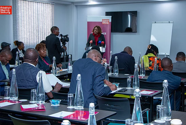 Conference room with attendees seated at tables, a woman speaking at a podium, and a cameraman recording the event.